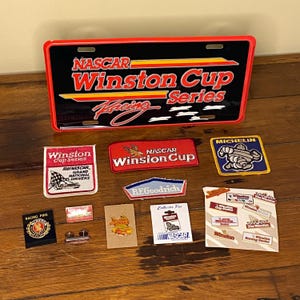 May include: A collection of NASCAR Winston Cup Series memorabilia, including a license plate, patches, and pins. The license plate is black with red and yellow accents. Patches feature logos like Michelin and BFGoodrich.