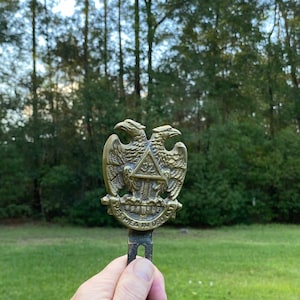 May include: A gold-toned metal Masonic emblem featuring two eagles with outstretched wings surrounding a triangle with the number "32" inside. The inscription "Spes Mea In Deo Est" is visible below the eagles.