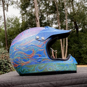 May include: A blue and purple motocross helmet with a black visor. The helmet features a swirling design in shades of blue, green, and red. The helmet is sitting on a dark surface with a forest background.