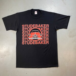 May include: Black t-shirt with a red and white graphic of a classic Studebaker car. The word "Studebaker" is stacked vertically in red letters on the left side of the car graphic.