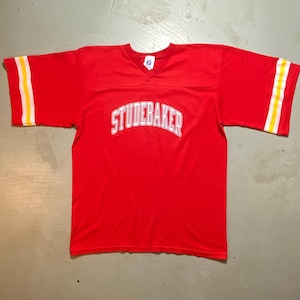 May include: A bright red short-sleeved jersey with a V-neck collar. The word STUDEBAKER is printed in white arched lettering across the chest. Each sleeve features two horizontal stripes in yellow and white near the hem. A white tag is visible.