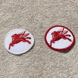 May include: Two embroidered patches featuring a red winged horse design on a white circular background. One patch has a white border, and the other has a red border. The patches are likely for clothing or accessories.