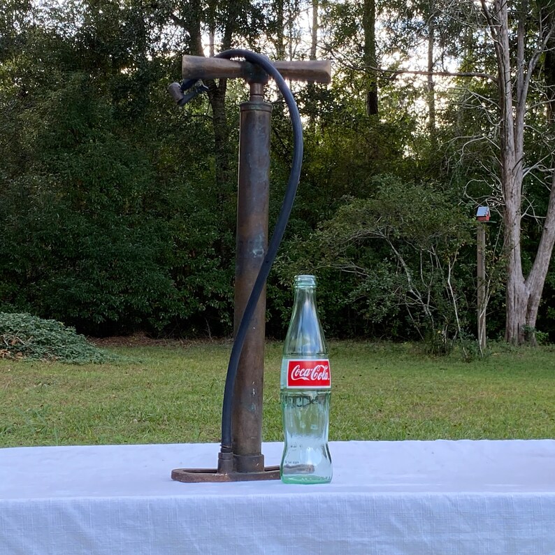 Puede incluir: Una bomba de bicicleta de lat&oacute;n vintage con una manguera de goma negra y una botella de Coca-Cola sobre un mantel blanco.