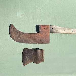 May include: A rusty, curved blade with a wooden handle and a separate rusty axe head. The tools are on a green surface.