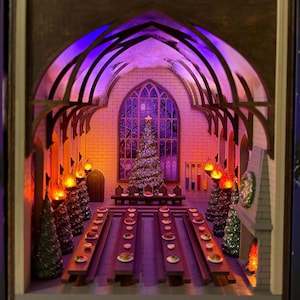 Könnte beinhalten: Ein hölzernes Diorama eines großen Saals mit Weihnachtsbaum, langen Tischen und gewölbten Decken. Die Szene wird von warmen und kühlen Lichtern beleuchtet, die eine festliche und magische Atmosphäre schaffen. Das Diorama ist von einem Holzrahmen umgeben.