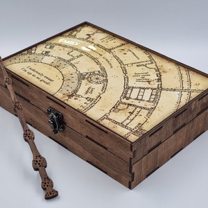 Puede incluir: Una caja de madera con un diseño de mapa en la tapa y el nombre "EMILY" impreso. Una varita está colocada al lado. El texto "I solemnly swear I'm up to no good..." está impreso en la caja. La caja tiene un cierre metálico.