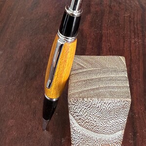 以下が含まれることがあります： 磨かれた仕上げの、手作りの木製ペン。シルバーのクリップとアクセントが付いています。ペンは、質感のある正方形の木製ブロックの隣に置かれています。ペンのバレルは暖かく金色で、ペンの先端は黒です。