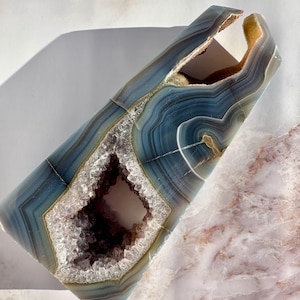 May include: A large, blue and brown agate geode with a natural opening revealing a crystal cave. The geode is shaped like a tall, thin pyramid.