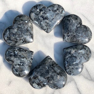 May include: Six polished heart-shaped stones made of blue and grey labradorite. The stones are arranged in a circle on a white marble surface.