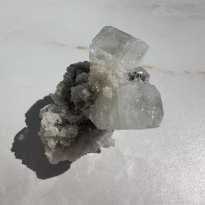 May include: A cluster of clear, crystalline minerals with a rough, textured base. The crystals are mostly rectangular and have a translucent appearance. The mineral formation is set against a white, marbled surface, with a dark shadow cast to the left.