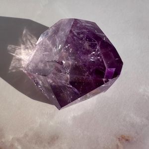 May include: A faceted amethyst crystal, displaying a rich purple colour. The crystal is cut into a geometric shape, reflecting light and creating shadows. The gemstone rests on a white and tan surface.