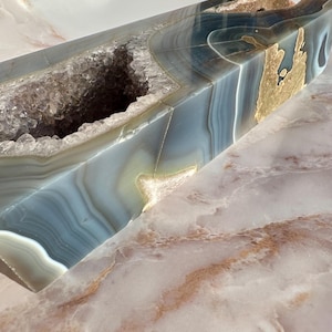 May include: A large, natural agate geode with a hollow center and a blue, gray, and gold color scheme. The geode is polished and has a smooth, shiny surface.