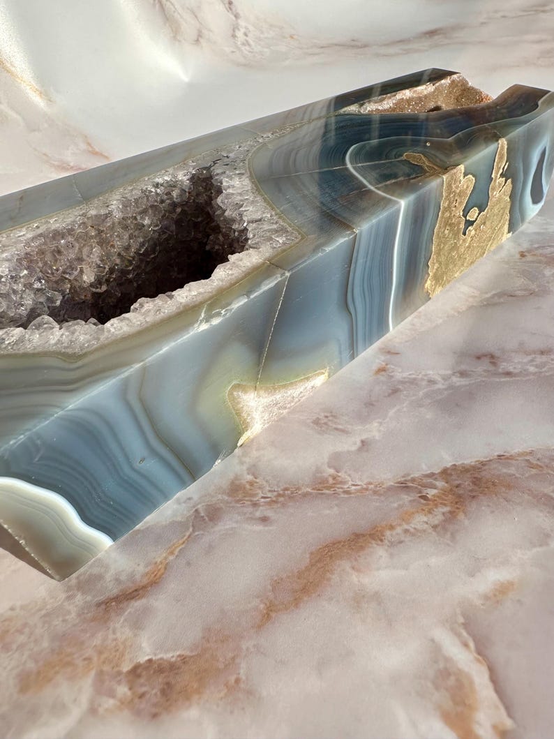 May include: A large, natural agate geode with a hollow center and a blue, gray, and gold color scheme. The geode is polished and has a smooth, shiny surface.