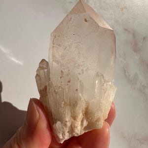 以下が含まれることがあります： 淡いピーチ色の透明な水晶。この結晶は、尖った上部と基部に小さな結晶のクラスターを持つ自然な形成物です。表面には自然な欠陥があります。