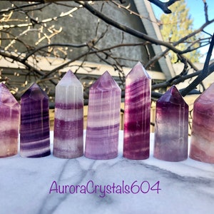 May include: Seven purple and white striped fluorite crystal points arranged in a row on a white marble surface. The crystals are all approximately the same size and shape, and they have a beautiful, iridescent sheen.