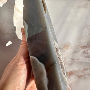 May include: A large, gray and white agate geode with a natural, rough edge. The geode has a large, open cavity that reveals the crystal formations inside.