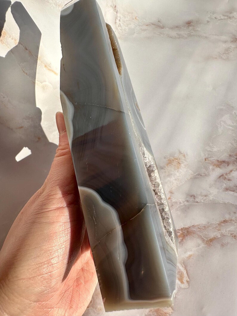 May include: A large, gray and white agate geode with a natural, rough edge. The geode has a large, open cavity that reveals the crystal formations inside.