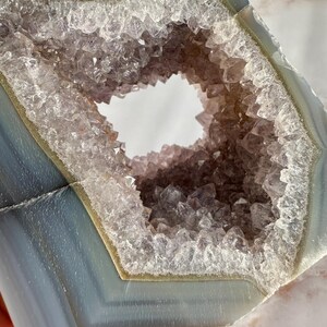May include: A large, natural agate geode with a hexagonal opening revealing a cluster of amethyst crystals. The geode is a light blue color with white and green banding.