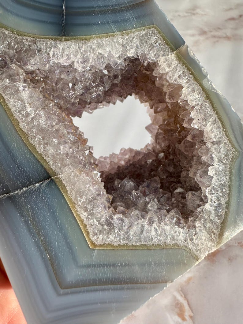 May include: A large, natural agate geode with a hexagonal opening revealing a cluster of amethyst crystals. The geode is a light blue color with white and green banding.