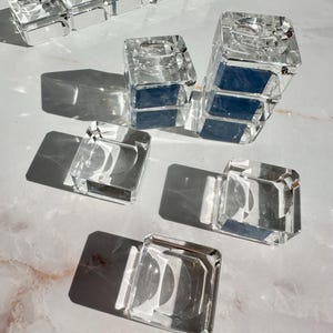 Pode incluir: Uma coleção de castiçais quadrados de vidro transparente com design facetado. O conjunto inclui várias peças individuais dispostas em grupo, com um centro oco para velas e uma base espessa e transparente que reflete a luz.