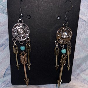 以下が含まれることがあります： シルバーとゴールドトーンのイヤリングのペア。各イヤリングは、中央にクリアストーン、ターコイズビーズ、ぶら下がっている矢のチャームが付いた円形のペンダントが特徴です。イヤリングは黒いカードに表示されています。