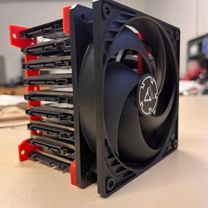 Puede incluir: Una carcasa de disco duro negra con un marco rojo y un ventilador negro con un logotipo Arctic blanco. La carcasa está apilada con varios discos duros.