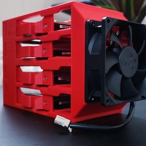 Puede incluir: Una carcasa roja para disco duro de ordenador con cuatro bahías. Un ventilador negro está montado en el lateral. Un conector blanco y un cable negro se extienden desde la carcasa. Diseñado para alojar y enfriar discos duros.