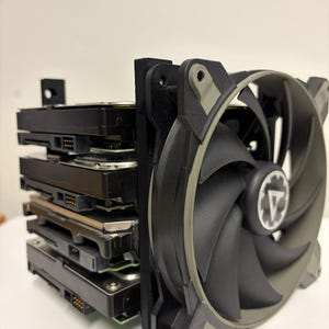 May include: A stack of four black hard disk drives mounted together with a large black and grey computer cooling fan attached to the front. The fan features a central circular logo with a white triangle and gear icon on a black background.