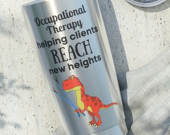May include: A stainless steel tumbler with a white lid. The tumbler features the text "Occupational Therapy helping clients REACH new heights" and a cartoon dinosaur illustration in red and yellow.