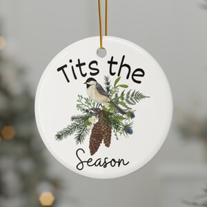 Może przedstawiać: Biała ceramiczna ozdoba choinkowa z czarno-białym ptakiem siedzącym na gałęzi z szyszkami i zielenią. Ozdoba ma napis "Tits the Season".
