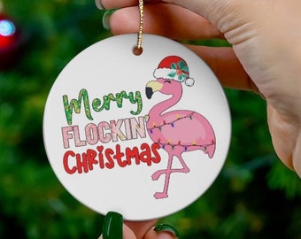 Może przedstawiać: Biała ceramiczna ozdoba choinkowa z różowym flamingiem w czerwonym kapeluszu Świętego Mikołaja i światełkami choinkowymi. Ozdoba ma napis "Merry Flockin' Christmas".