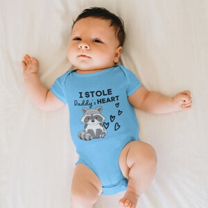 K&ouml;nnte beinhalten: Ein Baby tr&auml;gt einen hellblauen Strampler mit einem Waschb&auml;r-Motiv und dem Text "Ich habe Papas Herz gestohlen".