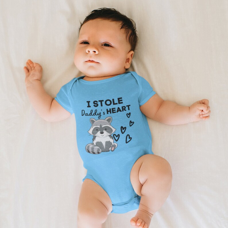 K&ouml;nnte beinhalten: Ein Baby tr&auml;gt einen hellblauen Strampler mit einem Waschb&auml;r-Motiv und dem Text "Ich habe Papas Herz gestohlen".