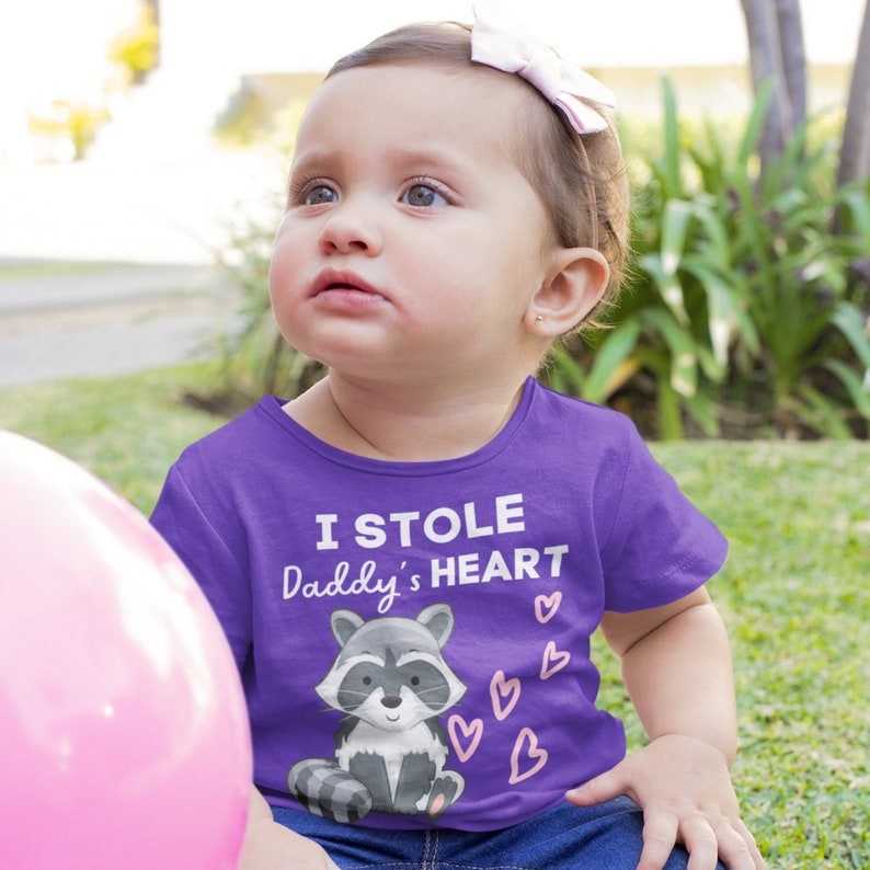 K&ouml;nnte beinhalten: Ein lilafarbenes T-Shirt f&uuml;r Kleinkinder mit einem Waschb&auml;r-Motiv und dem Text "Ich habe Papas Herz gestohlen".