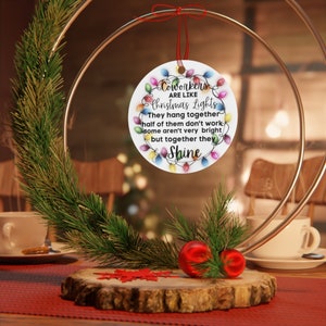 Puede incluir: Un adorno redondo blanco con una cadena de luces navide&ntilde;as de colores alrededor del borde. El adorno tiene el texto "Coworkers are like Christmas Lights. They hang together, half of them don't work, some aren't very bright, but together they Shine."