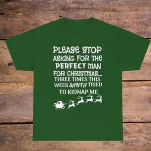 Pode incluir: Camiseta verde com texto branco que diz "PLEASE STOP ASKING FOR THE PERFECT MAN FOR CHRISTMAS... THREE TIMES THIS WEEK SANTA TRIED TO KIDNAP ME". Há cinco renas brancas puxando um trenó com o Papai Noel.