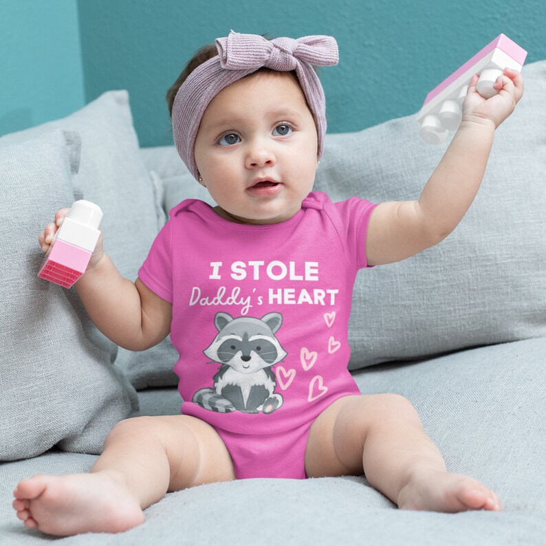 K&ouml;nnte beinhalten: Ein rosa Baby-Strampler mit kurzen &Auml;rmeln zeigt einen Cartoon-Waschb&auml;ren und den Text "I STOLE Daddy's HEART" in Wei&szlig;. Um den Waschb&auml;ren herum sind rosa Herzen verteilt.