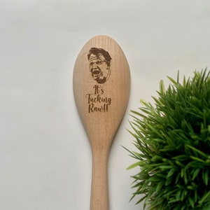 May include: A light brown wooden spoon with a portrait and the text "It's Fucking Raw!!" etched into the spoon. The spoon has a long handle. The text and portrait are dark brown.