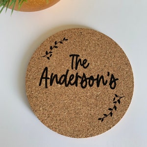 May include: A round cork coaster with the text "The Anderson's" in black cursive lettering. The coaster has a simple black floral design on the top and bottom.