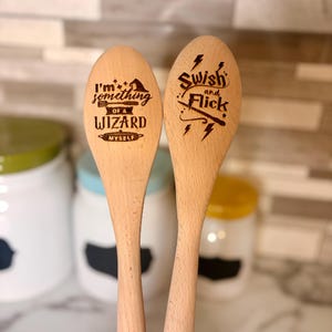May include: Two wooden spoons with engraved text. One spoon reads "I'm something of a WIZARD myself" and the other says "Swish and Flick". The spoons are light brown and have a smooth finish. The background is blurred.