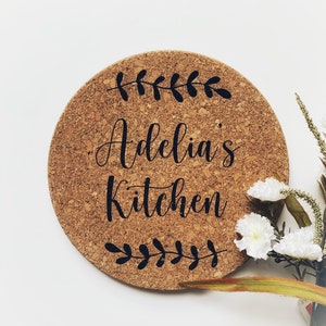Peut inclure: Un dessous de plat rond en liège avec le texte "Adelia's Kitchen" en lettres d'écriture noires. Le dessous de plat est décoré d'un simple motif de feuilles noires autour du bord.