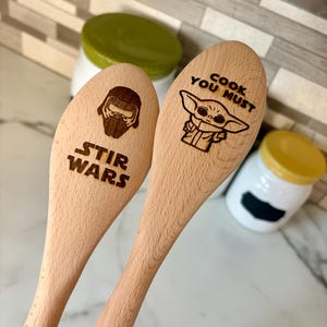 May include: Two wooden cooking spoons with engraved Star Wars designs. One spoon has "STIR WARS" and a Kylo Ren helmet. The other spoon has "COOK YOU MUST" and a Baby Yoda illustration.