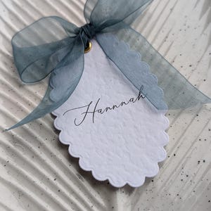 May include: A white scalloped tag with a textured surface featuring the name Hannah written in black cursive script. The tag is adorned with a translucent dusty blue organza ribbon tied in a bow through a small gold-toned metal eyelet at the top.
