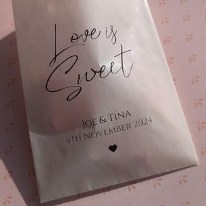 May include: White favour bag with the text "Love is Sweet" in black cursive font. Below the text is "Joe & Tina" and "6th November 2024" in black font. A small black heart is below the date.