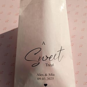 May include: A white paper bag with a clear window, featuring the text "A Sweet Treat" in black cursive font. Below the text, it reads "Alex & Mia 09.03.2025" with a small black heart symbol.