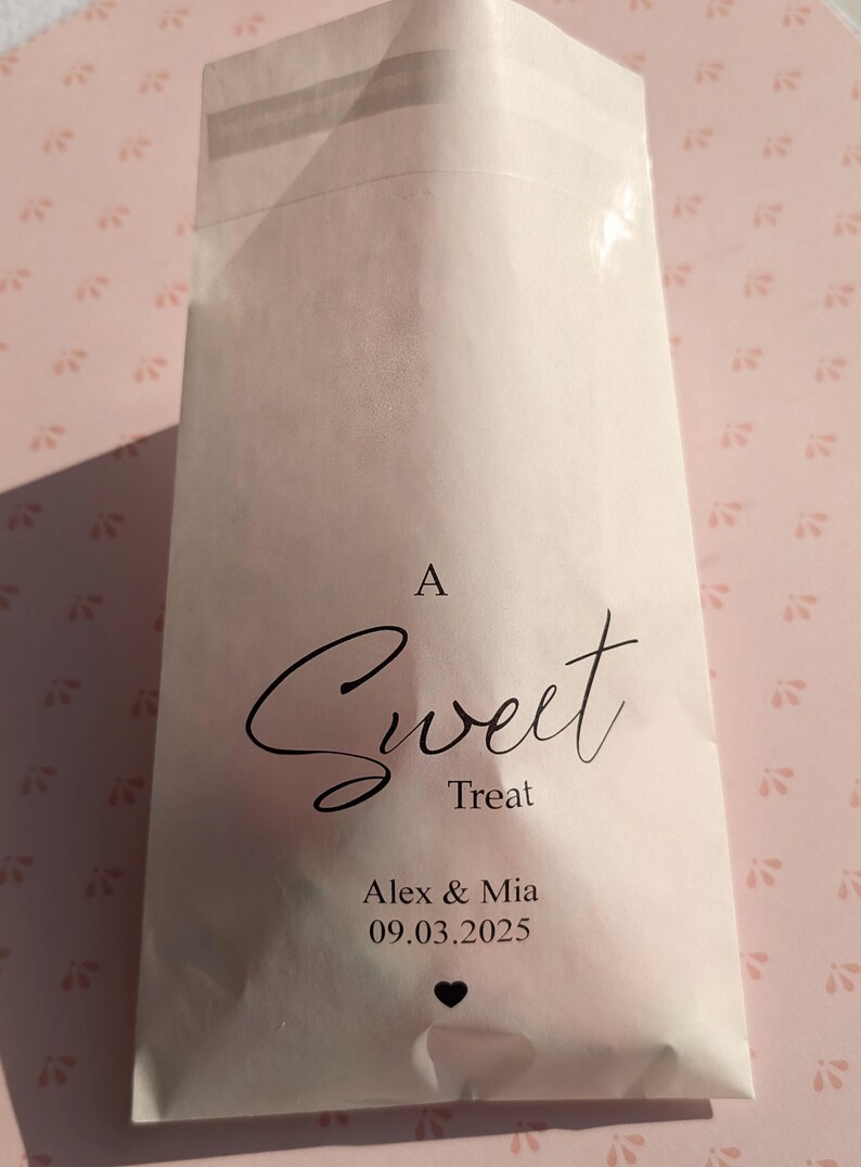 May include: A white paper bag with a clear window, featuring the text "A Sweet Treat" in black cursive font. Below the text, it reads "Alex & Mia 09.03.2025" with a small black heart symbol.