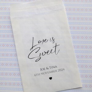 May include: White paper favour bag with the text "Love is Sweet" in black cursive script. Below the text is "Joe & Tina" and "6th November 2024" in black text. A small black heart is below the date.