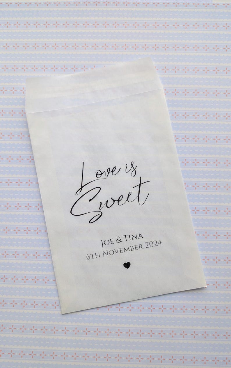 May include: White paper favour bag with the text "Love is Sweet" in black cursive script. Below the text is "Joe & Tina" and "6th November 2024" in black text. A small black heart is below the date.