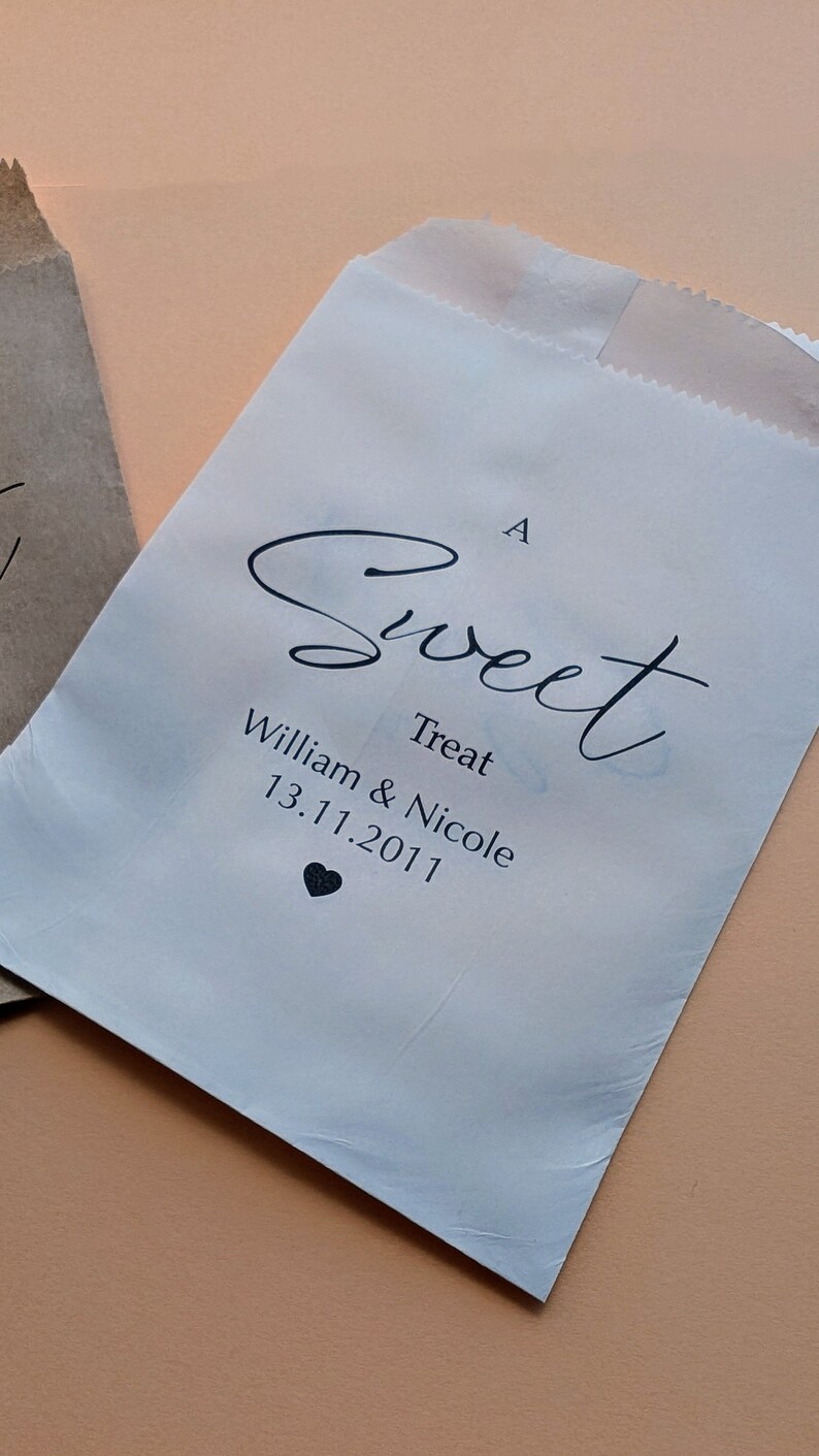 May include: A white paper bag with a black cursive inscription that reads "A Sweet Treat William & Nicole 13.11.2011" with a small black heart symbol below the date.