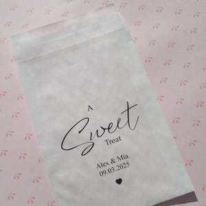 May include: A white paper bag with the text "A Sweet Treat" in black cursive font. Below the text is "Alex & Mia" and the date "09.03.2025" in black font. A small black heart is below the date.